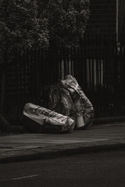 A pile of mixed waste and packaging materials, including cardboard boxes, paper bags, and plastic wrapping, is situated on the ground near a rough brick wall and the trunk of a tree. The cardboard box