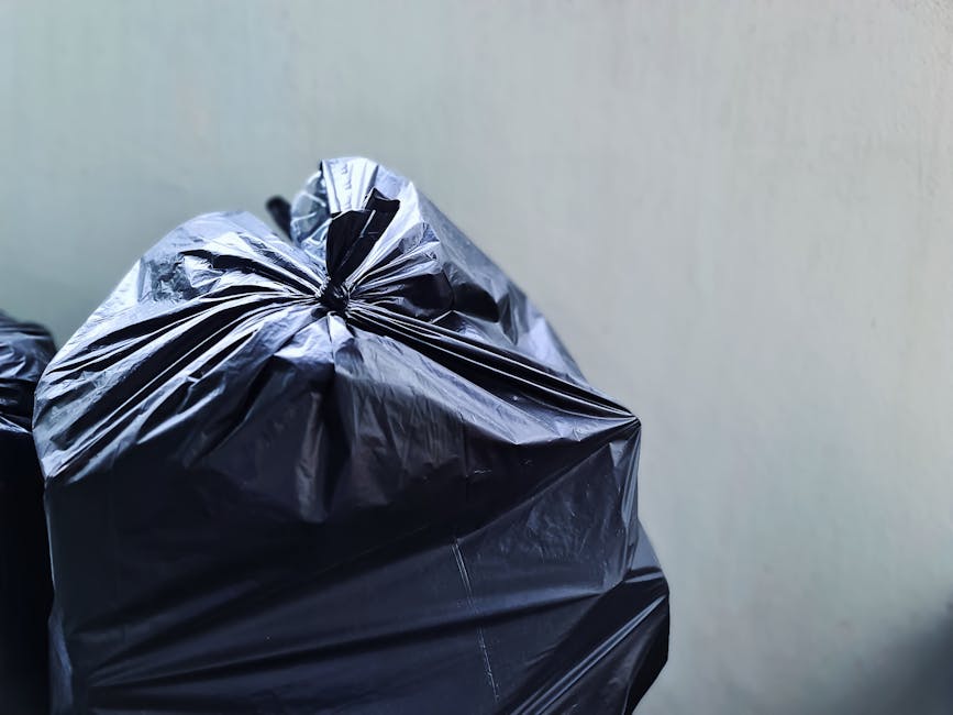 Close-up of black plastic rubbish bags filled with waste, stacked against a plain light-colored wall. The glossy surface of the bags reflects light, revealing crinkled textures and tightly knotted top